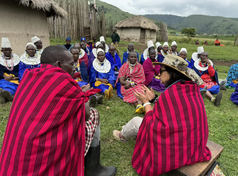 Maasai elders in red shukas and beaded necklaces sit in a circle on grassy hillside with huts and mountains, conversing with woman in hat.