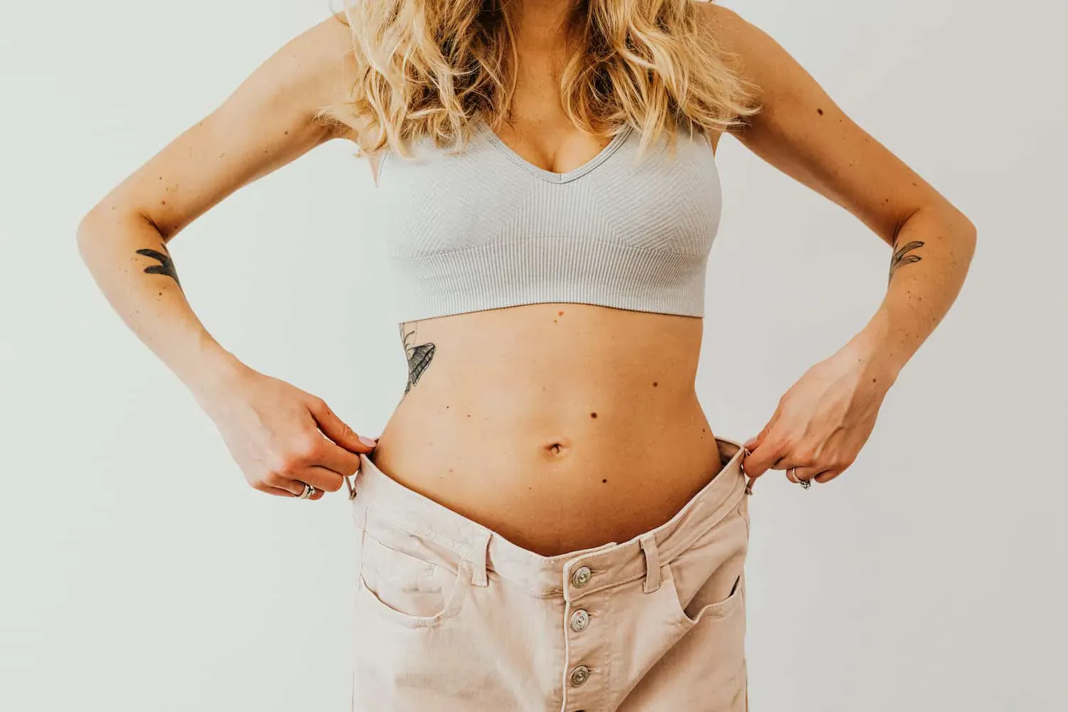 Blonde woman in gray sports bra and beige jeans, pulling waistband to show toned abs and tattoos
