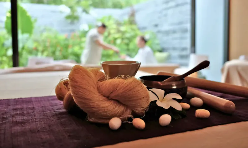 Spa table with orange scrub, frangipani flowers, eggs, ceramic bowls, spoons, and rollers; blurred therapists and garden beyond