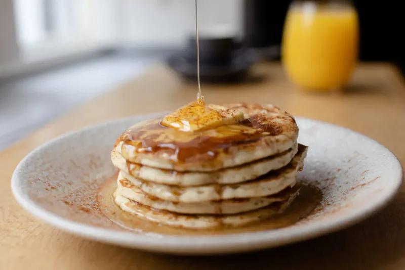 Stack of pancakes topped with butter and pouring maple syrup, beside orange juice on a table.