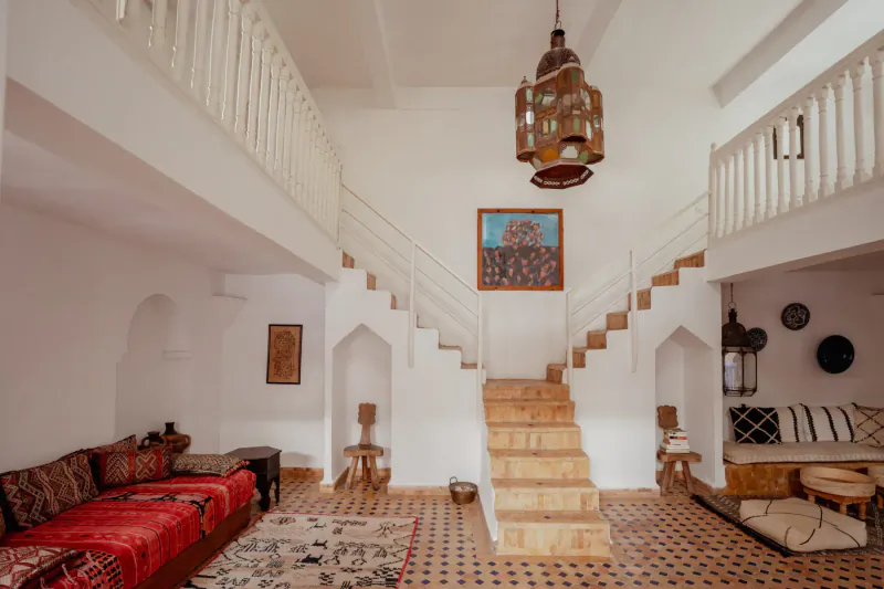 Moroccan villa living room with red cushions, ornate lantern, wooden stairs to white balcony, tiled floor, Essaouira luxury.