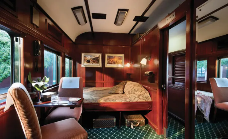 Luxurious wooden Rovos Rail train cabin with double bed, armchairs, desk, lamps, and green carpet.