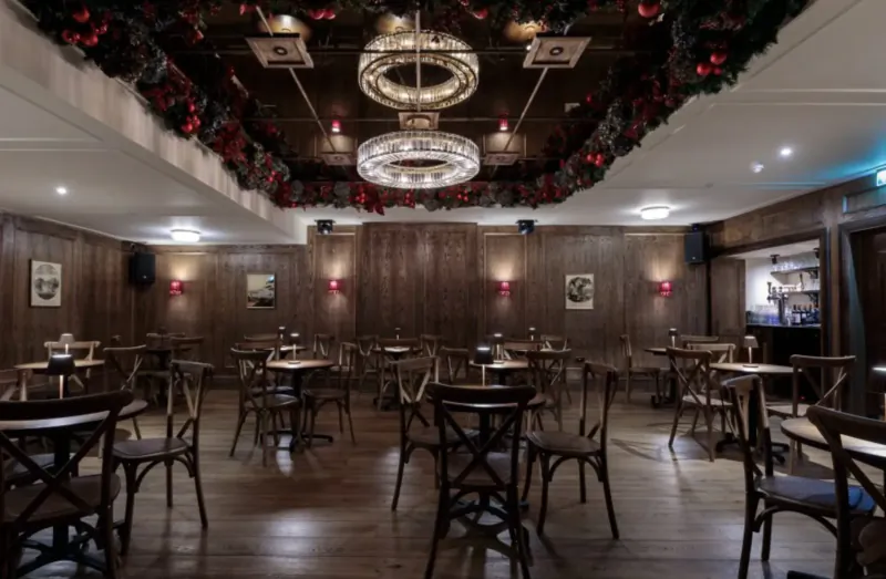 Festive interior of The Trafalgar Public House gallery with garland ceiling, chandelier, wood-paneled walls, and round tables with chairs.