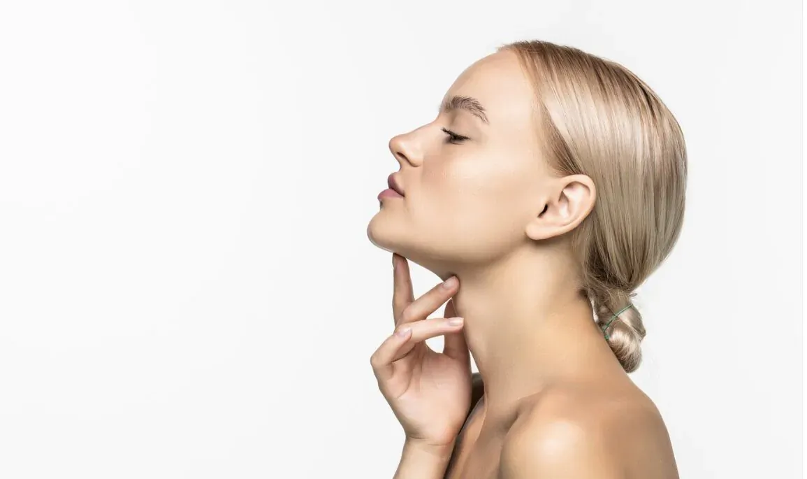 Blonde woman in profile touching neck with fingers, demonstrating face sculpting massager pose on white background
