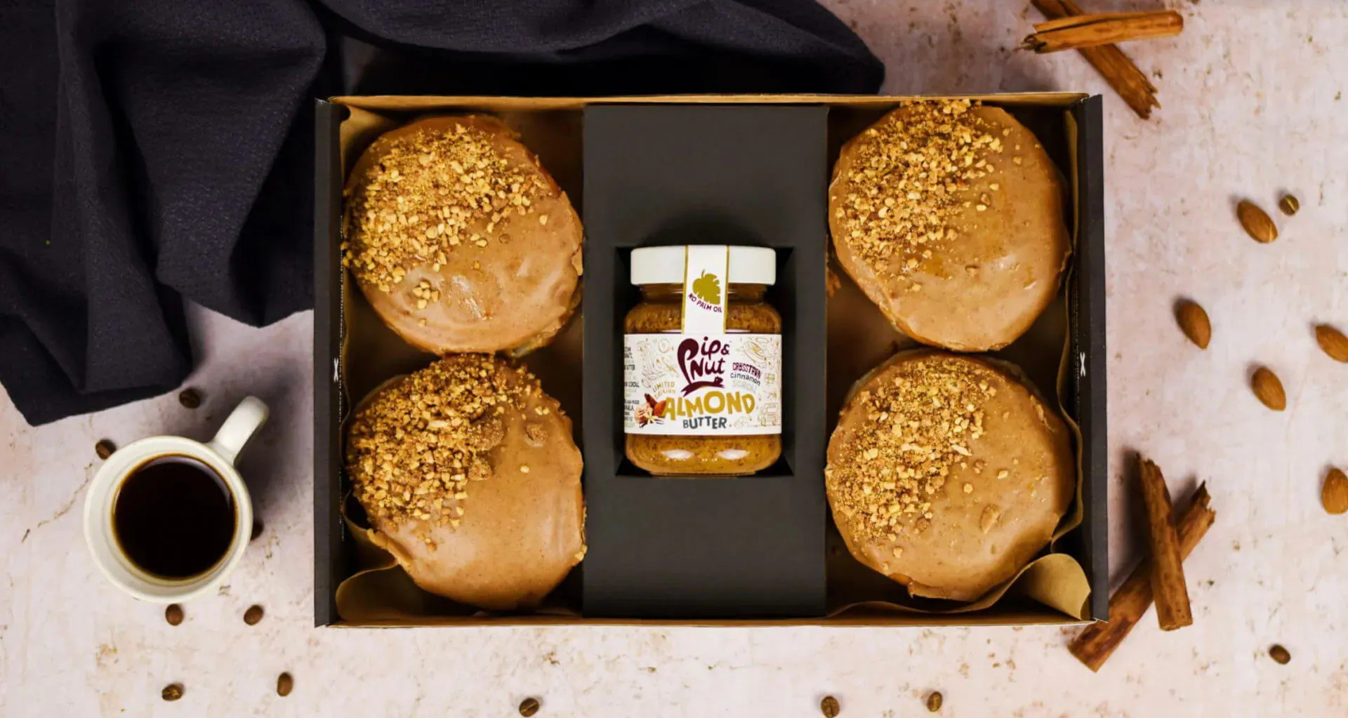 Open black box with 4 vegan Crosstown doughnuts topped with Pip & Nut almond butter, cinnamon sticks, almonds, and coffee cup.