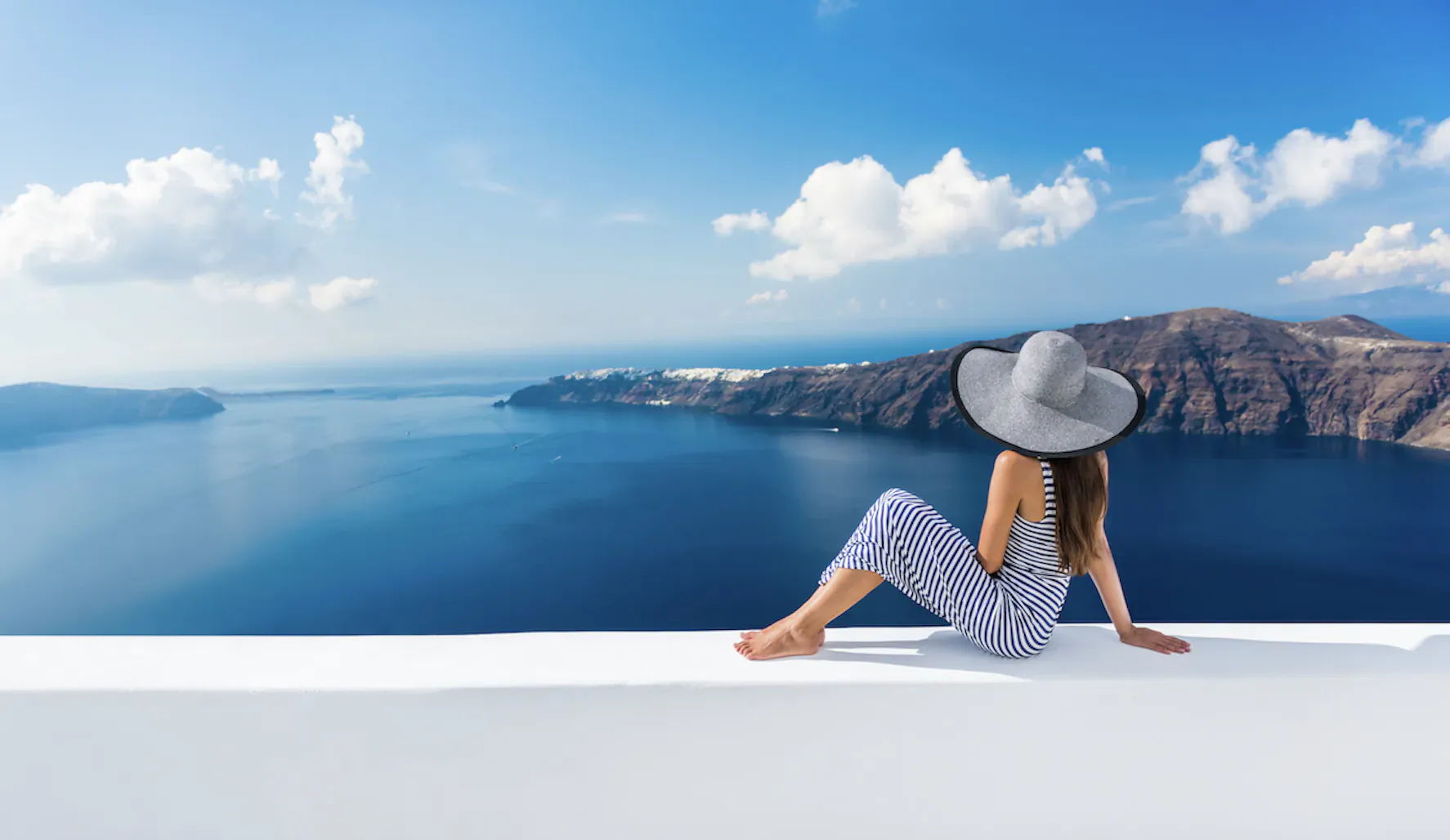 Woman in gray floppy hat and blue dress sits barefoot on white ledge, gazing at Santorini's blue caldera and cliffs under clear sky