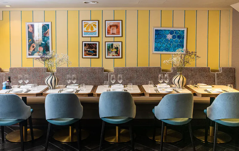 Elegant Bellazul restaurant dining area with yellow striped walls, abstract art, set table, and blue velvet chairs.