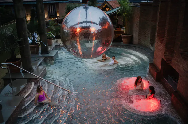 Large glowing silver orb over pool at Rabbit Hotel retreat; women relax in hot tubs and swim in stylish indoor spa.