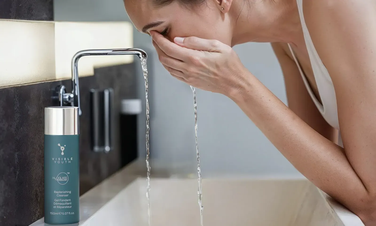 Woman splashing water on face at sink with Visible Youth Replenishing Cleanser bottle nearby