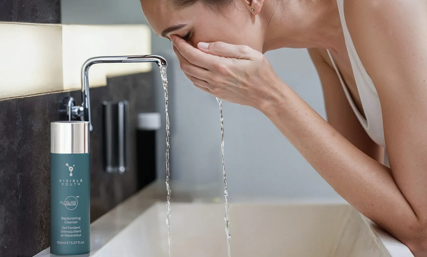 Woman splashing water on face at sink with Visible Youth Replenishing Cleanser bottle nearby