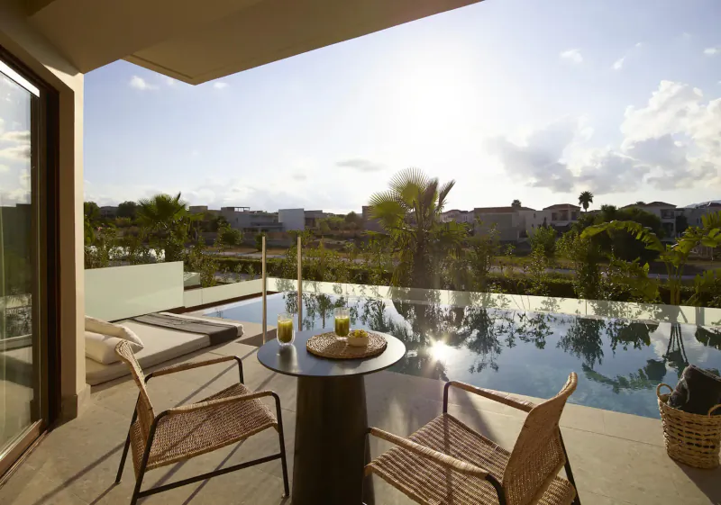 Sunny terrace at Asterion Suites & Spa, Crete, with infinity pool, lounge chairs, round table with drinks, palm trees, and sea view.