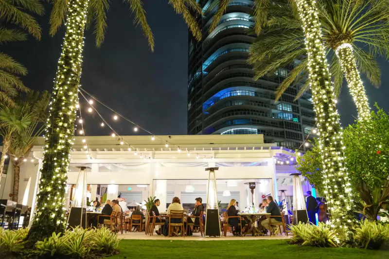 Nighttime outdoor dining at Continuum and K'Far with palm trees, string lights, curved high-rise, and patrons.