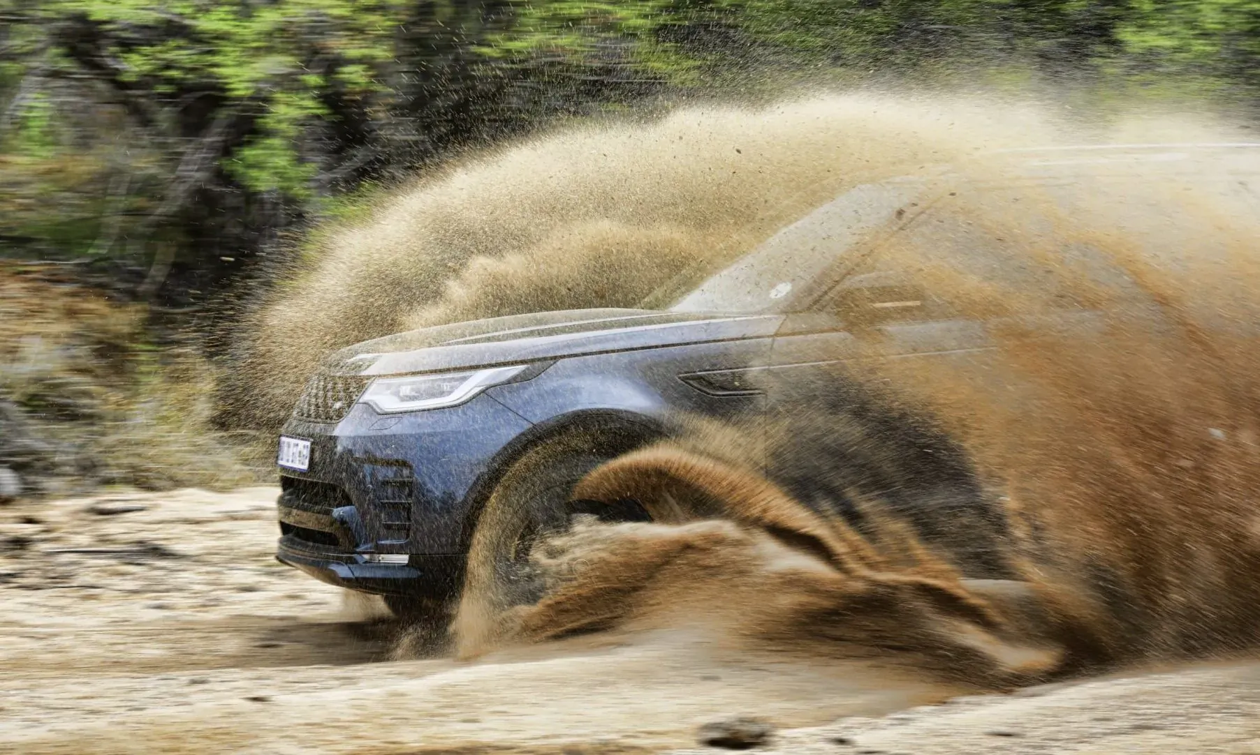 Blue Land Rover Discovery D350 HSE speeding through dusty off-road trail, kicking up clouds of dirt amid green trees.