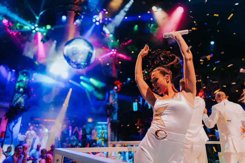 Singer in white outfit holds mic, dances energetically on stage with disco ball, colorful lights, confetti at Mama Mia! The Party show