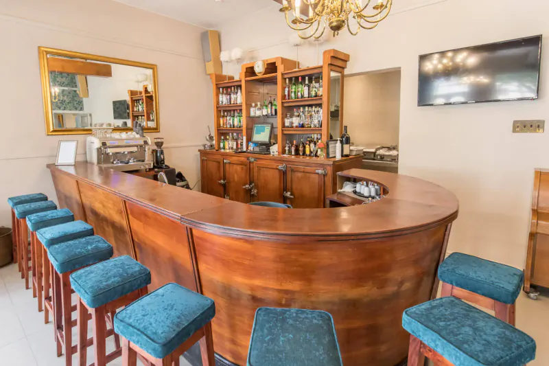 Curved wooden bar at Montagu Country Hotel with blue cushioned stools, shelves of bottles, mirror, and chandelier.