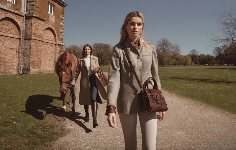 Two stylish women in tweed jackets and boots walk with bay horse on path by stone manor house in sunny park.