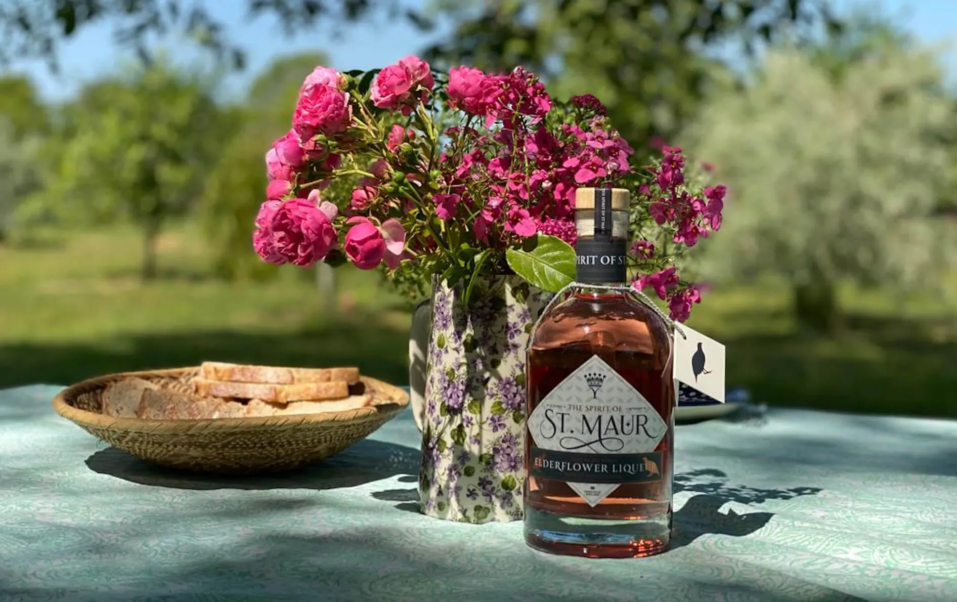 St. Maur tawny liqueur bottle on outdoor table with bread, pink roses, and green meadow backdrop