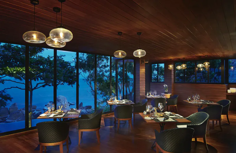 Elegant dining room at Silavadee Pool Spa Resort with wooden tables set for meals, pendant lights, and sunset sea view through large windows.
