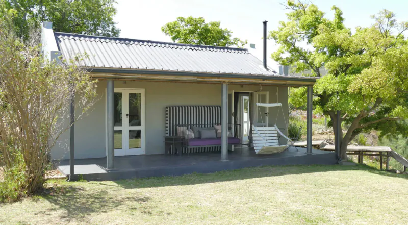 Grey cottage with verandah, purple bench, swing chair, and picnic table amid green trees and lawn at South Hill Vineyards, Elgin Valley.