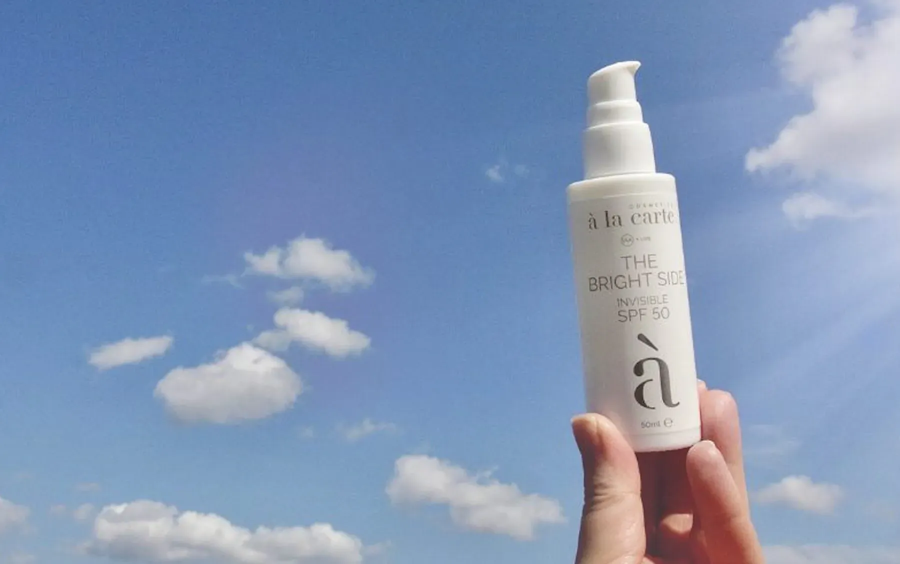 Hand holding white 'à la carte Launch the bright side invisible spf 50' sunscreen bottle against blue sky with clouds