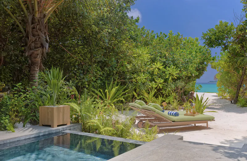 Luxurious Maldives villa poolside with green lounger, towel, tropical plants, white sand path to turquoise sea.