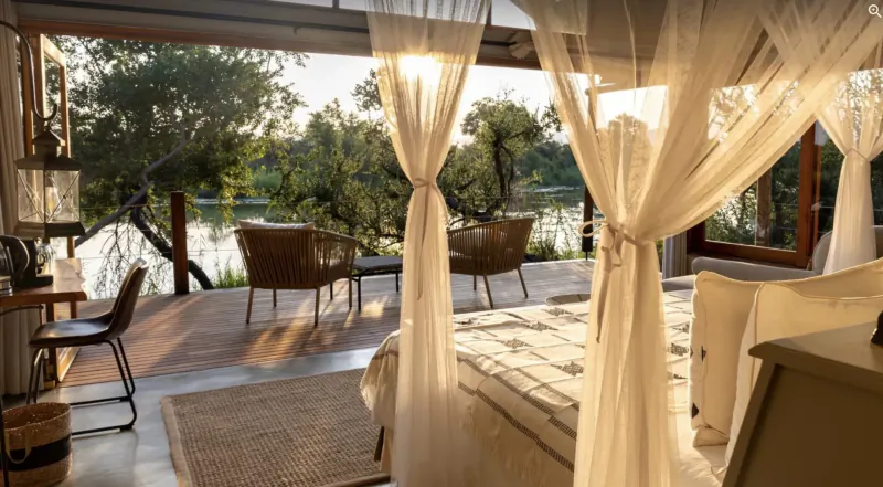 Luxurious open-air bedroom with white canopy drapes, rattan chairs, and deck overlooking river in African safari lodge