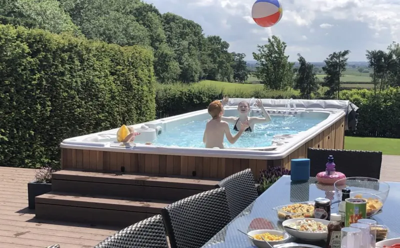 Boy holding baby in blue-lit hot tub on wooden deck, tossing beach ball, surrounded by hedges, fields, and outdoor dining table