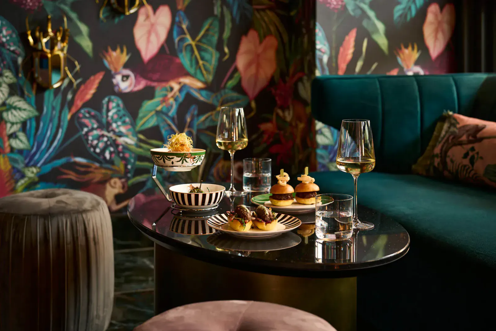 Luxurious lounge table with tropical wallpaper: white wine glasses, bowls of food, sliders on plates, emerald sofa, stools.