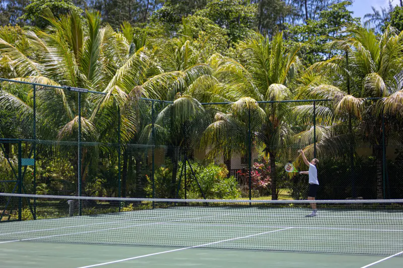 Person in white shirt playing tennis on green court at La Cigale Estate, surrounded by palm trees and fence, Seychelles.