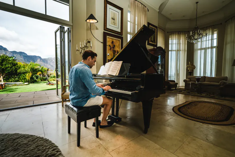 Man in blue shirt plays grand piano in luxurious room with mountain and garden view at 21 Nettleton, Cape Town.