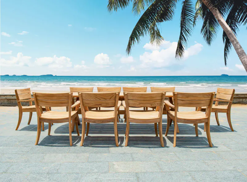 Teak rectangular dining table with 10 chairs on a terrace overlooking beach and palm trees under blue sky.