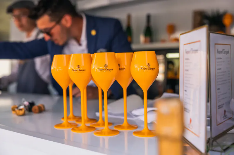 Row of orange Veuve Clicquot champagne glasses on white bar counter, bartenders in suits behind, event setting.