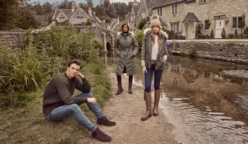 Man sitting and two women standing in stylish coats, boots by quaint stone cottages and stream.