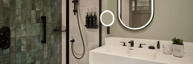 Modern bathroom with green tiled shower, white sink, round backlit mirror, toiletries, and plants