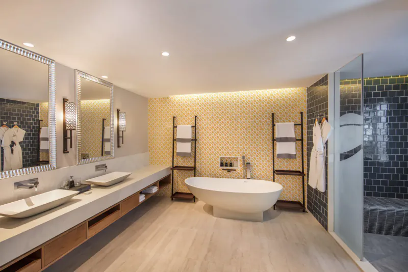 Luxurious hotel bathroom with freestanding white bathtub, double sinks, robes on racks, and glass shower at Anantara Iko Mauritius Resort.