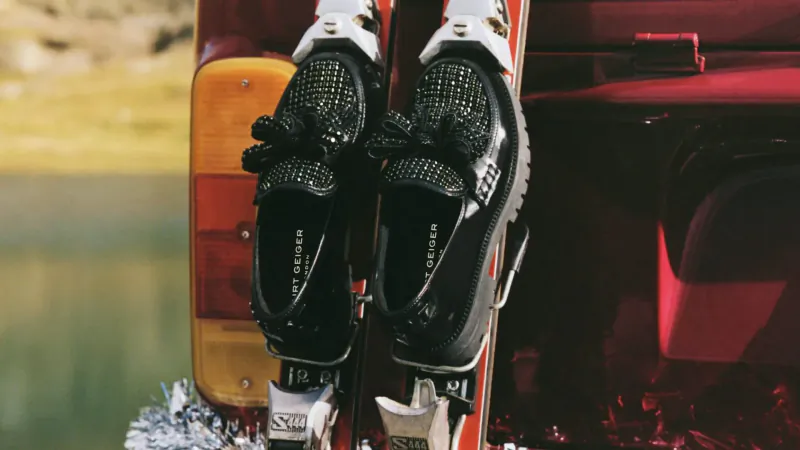 Close-up of black sparkly Kurt Geiger loafers strapped to white skis on red Jeep by lake, holiday vibe.