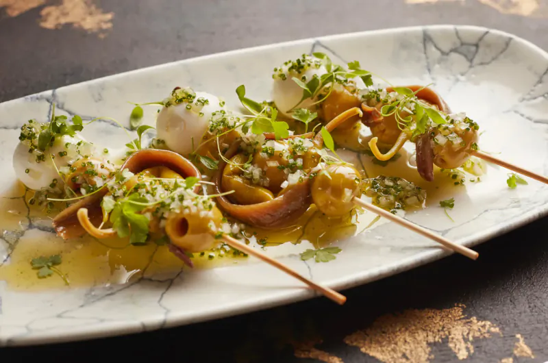 Four skewers of burrata, tuna tataki, and microgreens on a white marbled plate