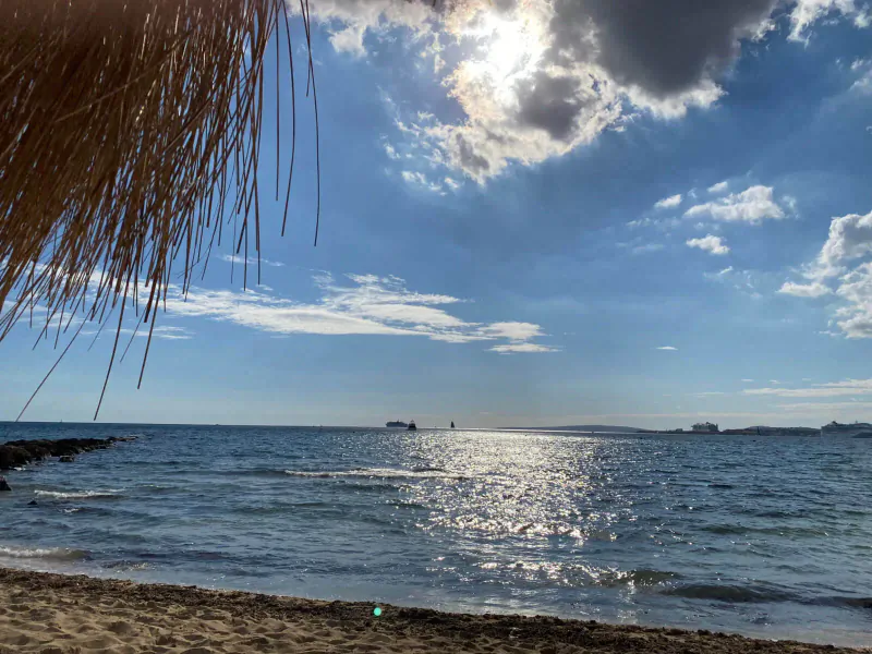Sunny beach view at Assaona Gastrobeach Palma Club with thatched umbrella, calm sea, distant ships, and clouds.