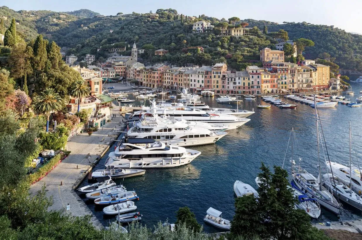 Aerial view of Portofino harbor with luxury white yachts docked amid colorful hillside buildings and lush greenery.