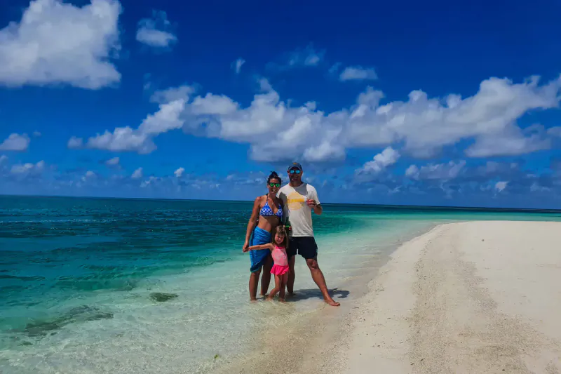 Family of four standing in shallow turquoise water at a pristine Seychelles beach, with white sandbar and blue sky.