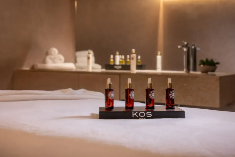 Four red KOS essential oil bottles on black stand on white massage bed in spa room with towels and plants