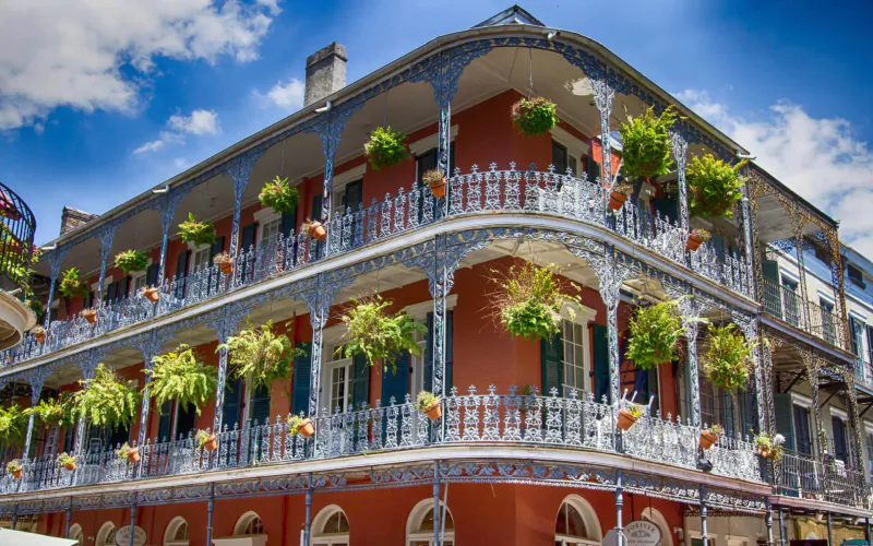 Corner red Creole townhouse in New Orleans with ornate iron balconies overflowing with green hanging plants under blue sky.