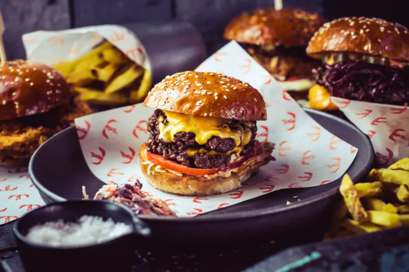 Close-up of a gourmet cheeseburger with beef patty, melted cheese, lettuce, tomato on a sesame bun, served on a plate with fries and red logo papers.