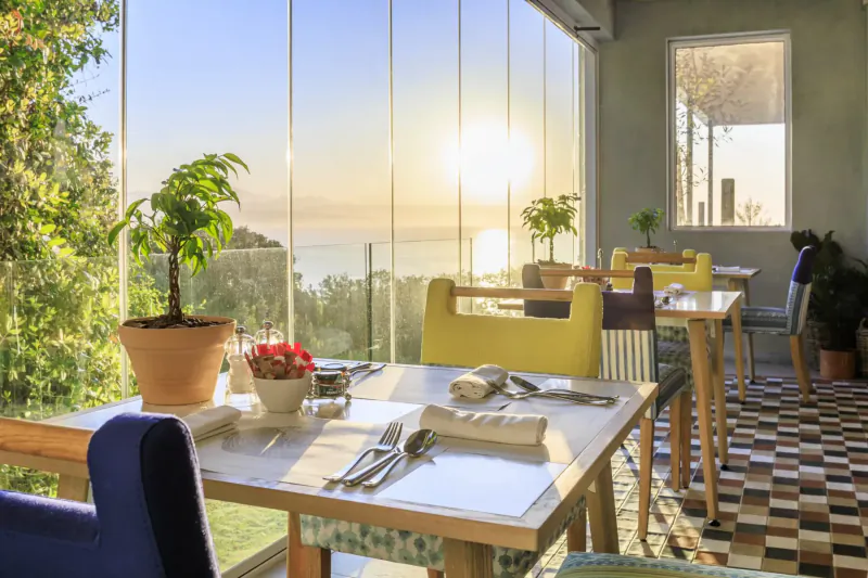 Sunset view over sea from The Plettenberg's elegant dining area with set tables, yellow chairs, and potted plants.