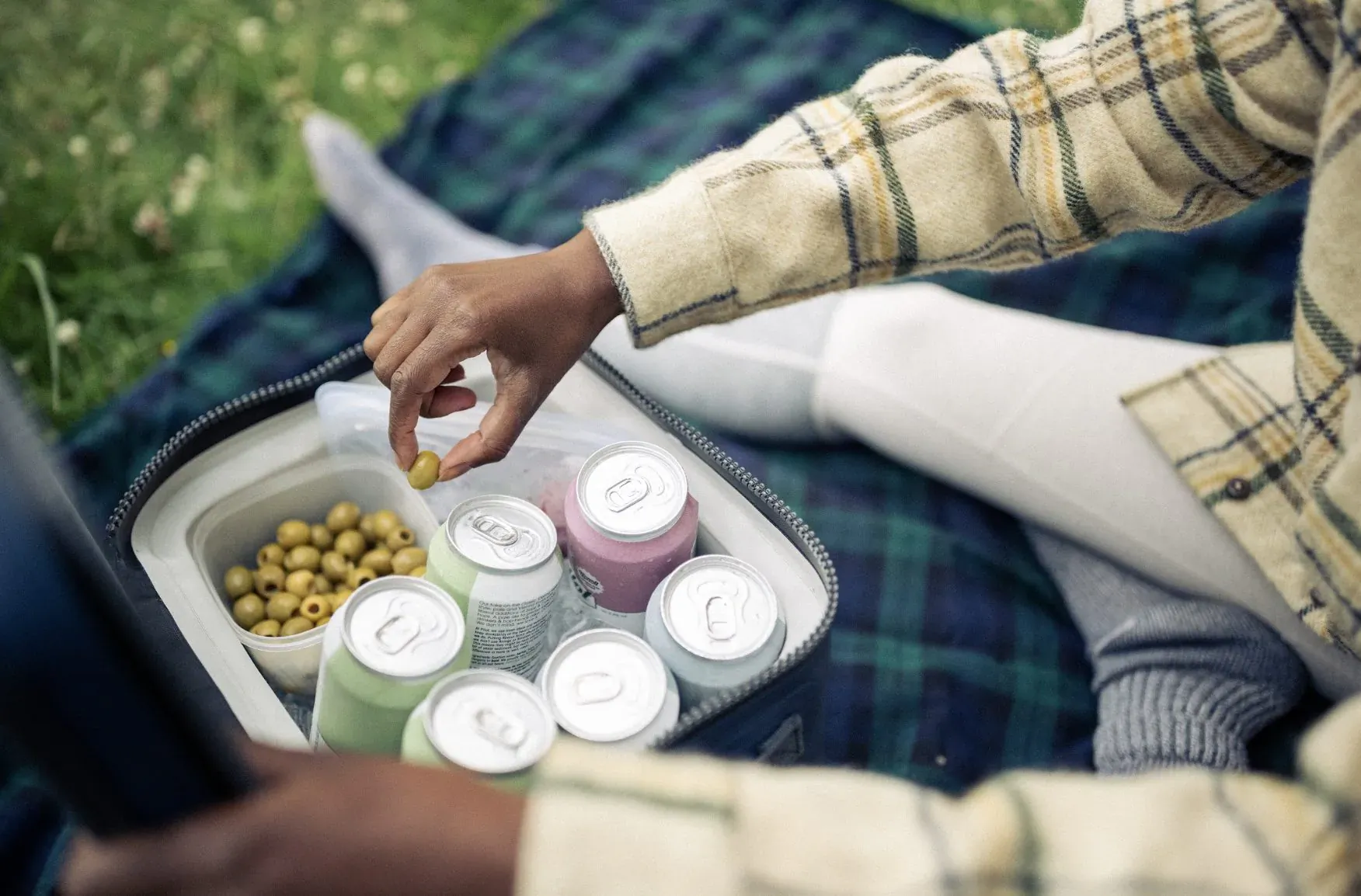 Person on picnic blanket picking green olives from cooler with colorful cans, wearing plaid jacket and white pants outdoors.