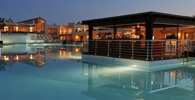 Twilight view of Cavo Spada resort: modern white buildings, elevated poolside bar, infinity pool with reflections.