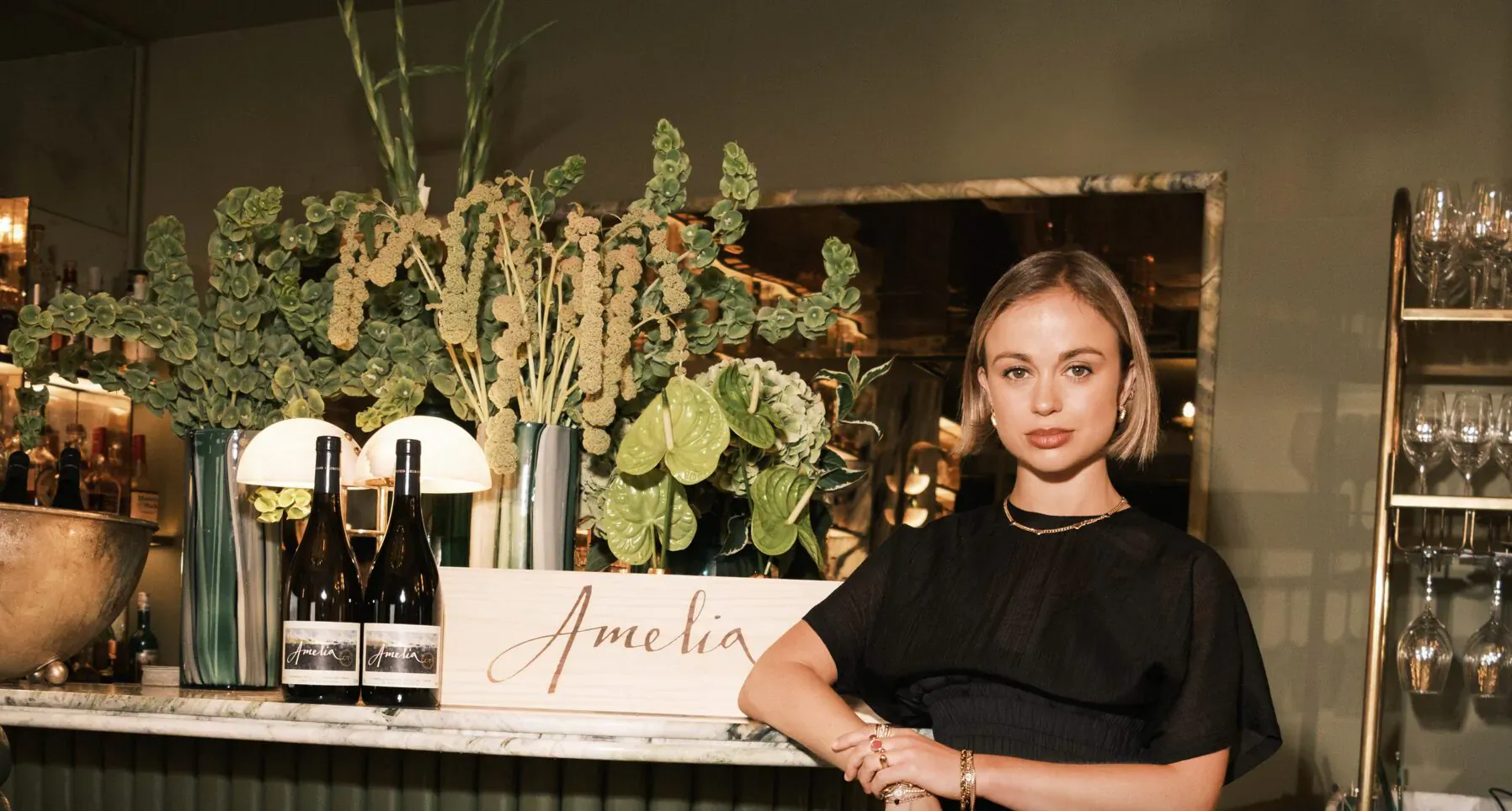 Lady Amelia Windsor smiling beside 'Amelia' wine bottles and lush green plants at her birthday lunch event