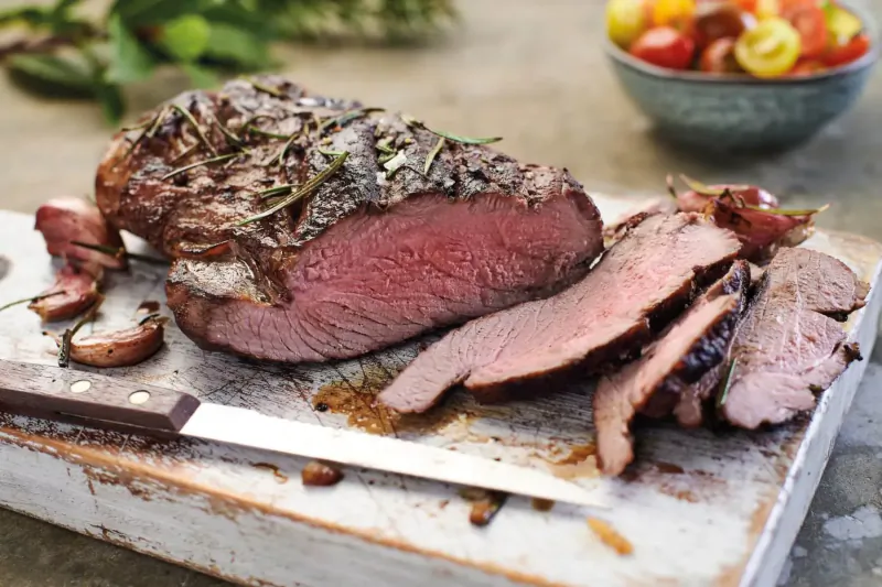 Sliced medium-rare roast beef with herbs on wooden board, knife, cherry tomatoes, and greens beside.