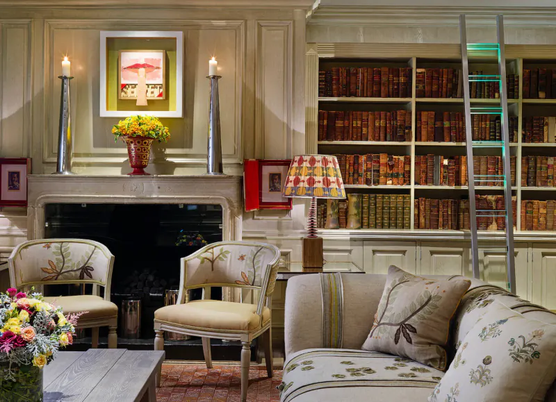 Elegant library room with bookshelves, ladder, floral armchairs, sofa, lit fireplace, candles, and yellow flowers.