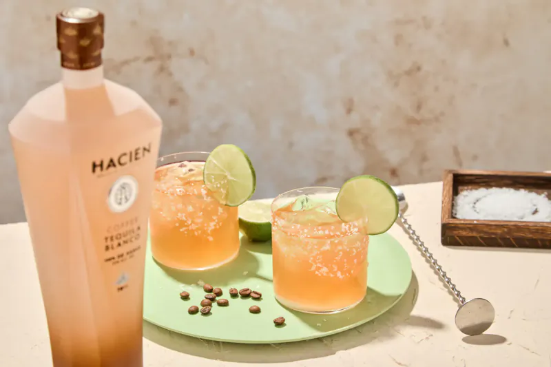 Hacienda pink tequila bottle beside two orange margaritas garnished with lime wedges and salt rim on green plate